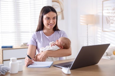 Photo of Telehealth. Smiling mother with her baby having online consultation with doctor and writing at table indoors
