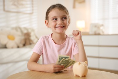 Photo of Little girl with pocket money and piggy bank at wooden table indoors