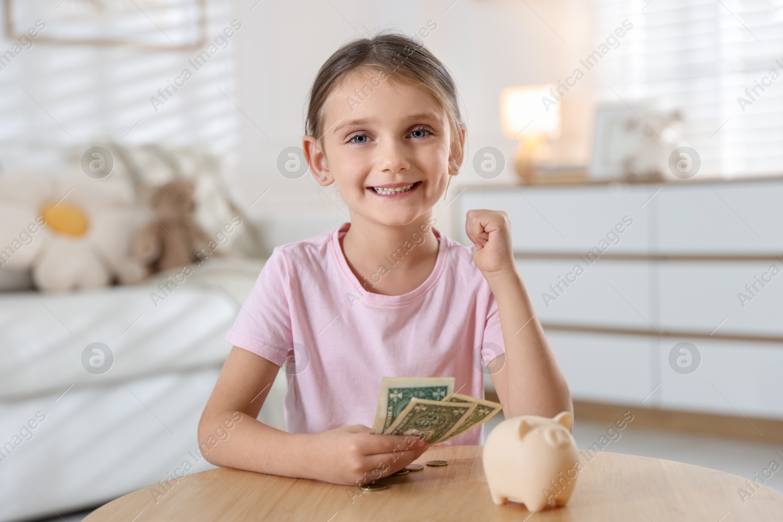 Photo of Little girl with pocket money and piggy bank at wooden table indoors