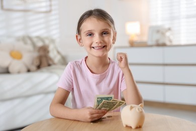 Photo of Little girl with pocket money and piggy bank at wooden table indoors