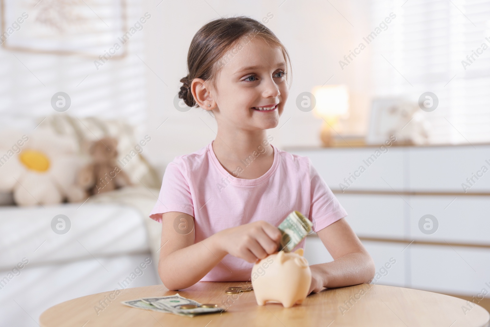 Photo of Little girl putting pocket money into piggy bank at wooden table indoors