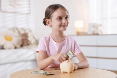 Photo of Little girl putting pocket money into piggy bank at wooden table indoors