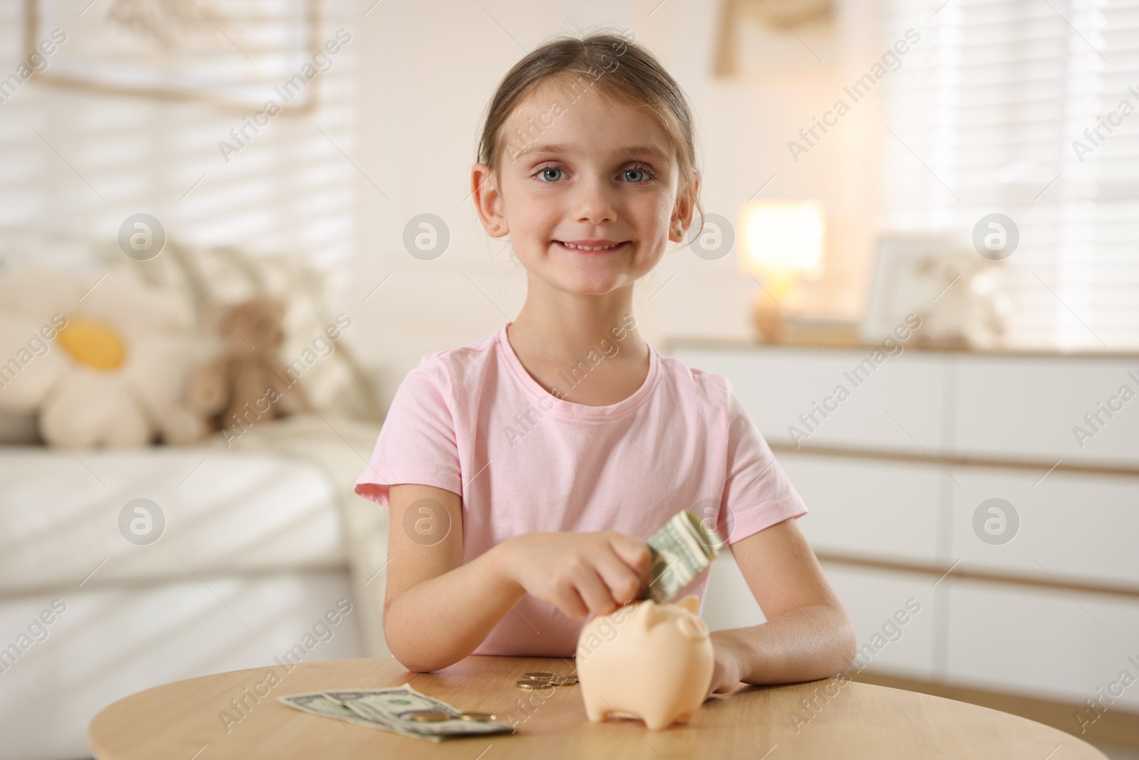 Photo of Little girl putting pocket money into piggy bank at wooden table indoors