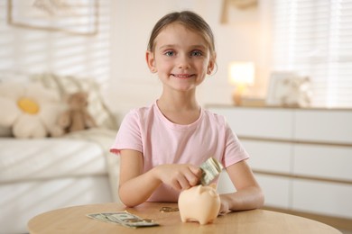 Photo of Little girl putting pocket money into piggy bank at wooden table indoors