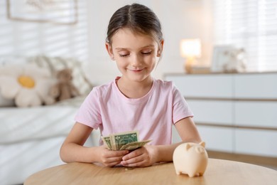 Photo of Little girl with pocket money and piggy bank at wooden table indoors