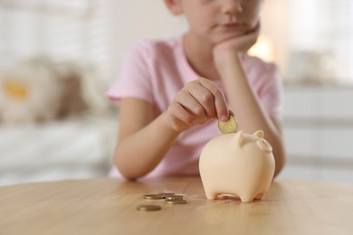 Photo of Little girl putting pocket money into piggy bank at wooden table indoors, closeup