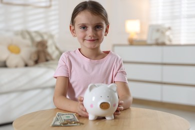 Photo of Little girl with pocket money and piggy bank at wooden table indoors