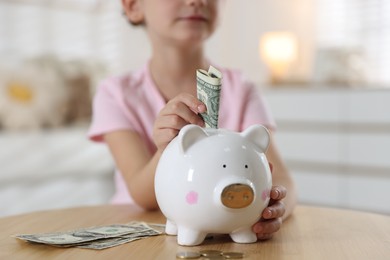 Photo of Little girl putting pocket money into piggy bank at wooden table indoors, closeup