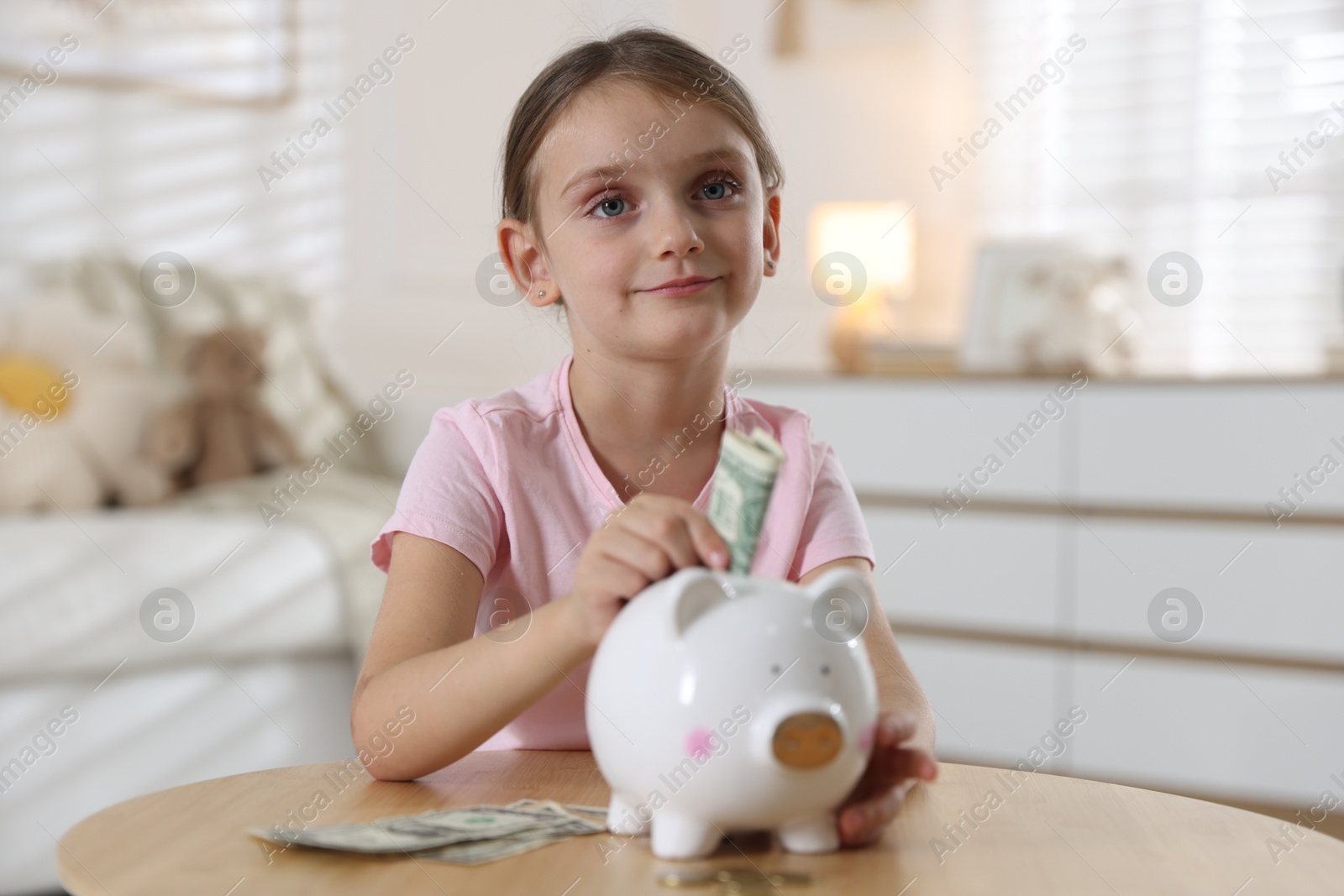 Photo of Little girl putting pocket money into piggy bank at wooden table indoors