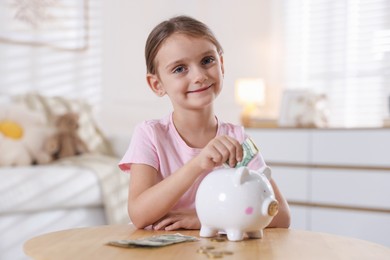 Photo of Little girl putting pocket money into piggy bank at wooden table indoors