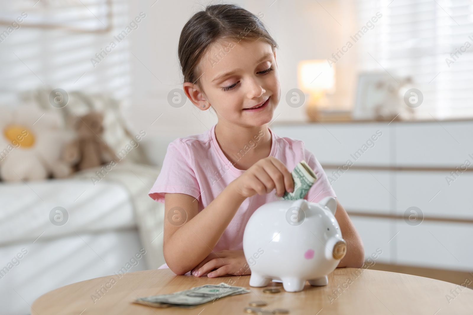 Photo of Little girl putting pocket money into piggy bank at wooden table indoors