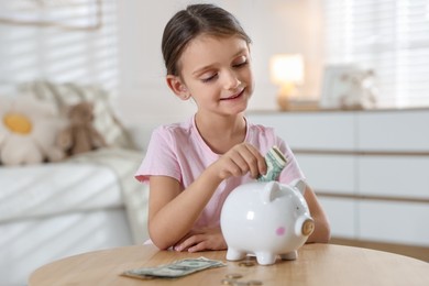 Photo of Little girl putting pocket money into piggy bank at wooden table indoors