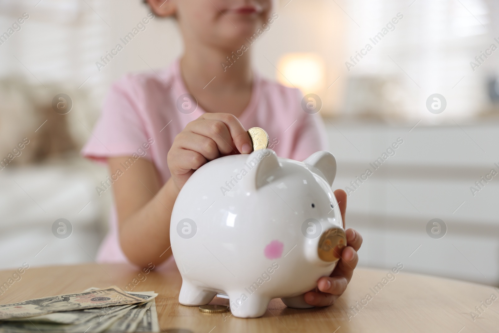 Photo of Little girl putting pocket money into piggy bank at wooden table indoors, closeup