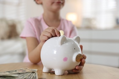 Photo of Little girl putting pocket money into piggy bank at wooden table indoors, closeup