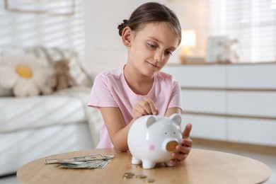 Photo of Little girl putting pocket money into piggy bank at wooden table indoors