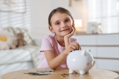 Photo of Little girl putting pocket money into piggy bank at wooden table indoors