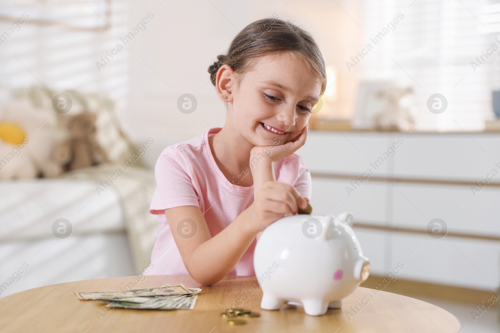 Photo of Little girl putting pocket money into piggy bank at wooden table indoors