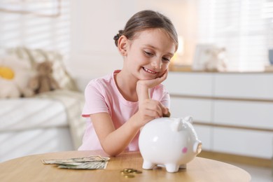 Photo of Little girl putting pocket money into piggy bank at wooden table indoors