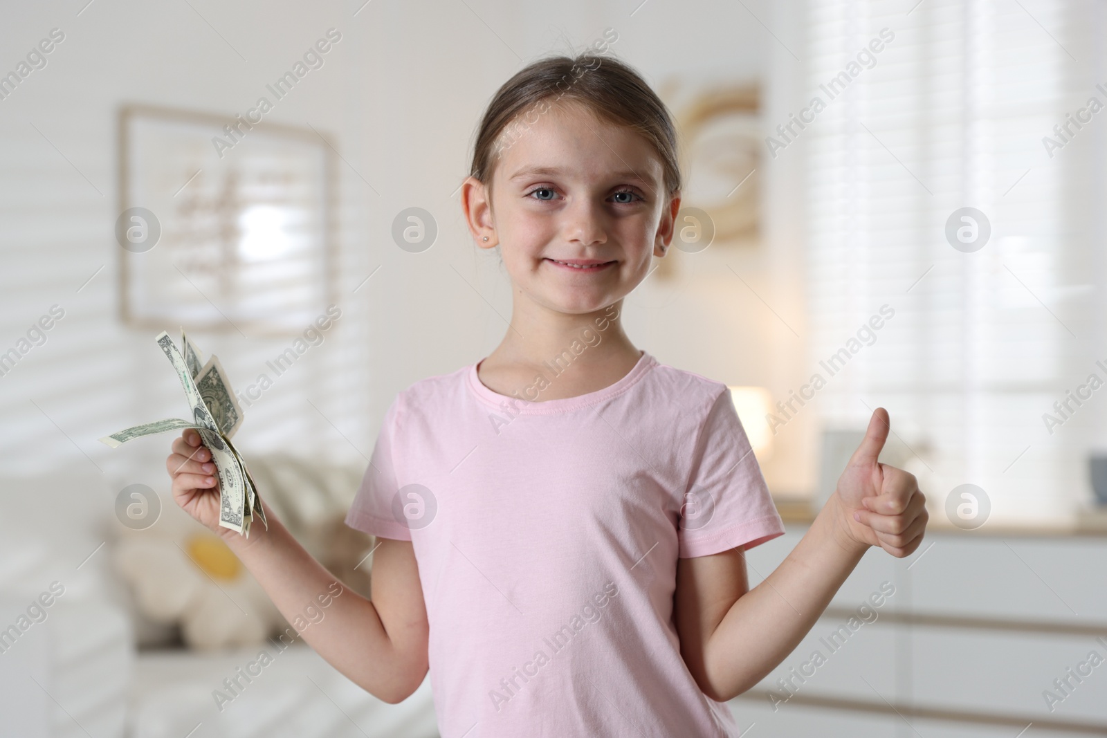 Photo of Little girl holding pocket money and showing thumbs up at home