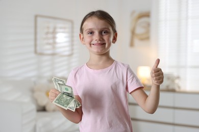 Photo of Little girl holding pocket money and showing thumbs up at home