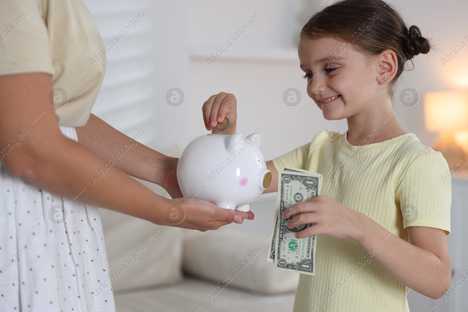 Photo of Mother and her daughter putting pocket money into piggy bank at home, closeup