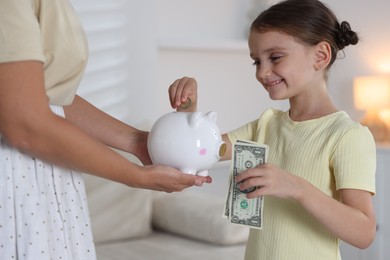 Photo of Mother and her daughter putting pocket money into piggy bank at home, closeup