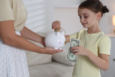 Photo of Mother and her daughter putting pocket money into piggy bank at home, closeup