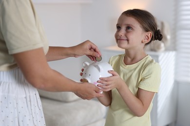 Photo of Mother and her daughter putting pocket money into piggy bank at home, closeup