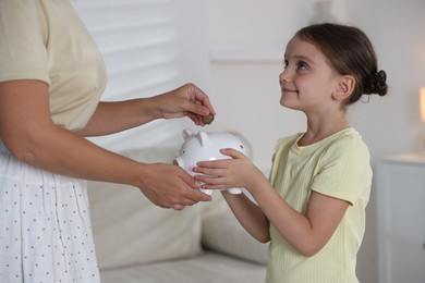 Photo of Mother and her daughter putting pocket money into piggy bank at home, closeup