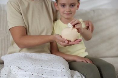Photo of Mother and her daughter putting pocket money into piggy bank at home, closeup