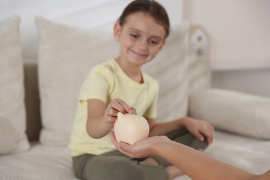 Photo of Mother and her daughter putting pocket money into piggy bank at home, closeup
