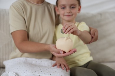 Photo of Mother and her daughter putting pocket money into piggy bank at home, closeup