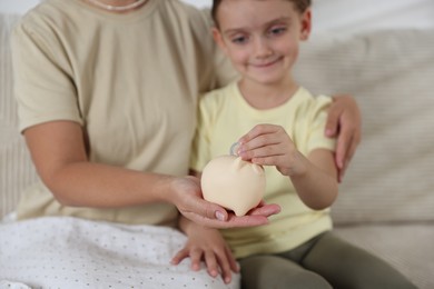 Photo of Mother and her daughter putting pocket money into piggy bank at home, closeup