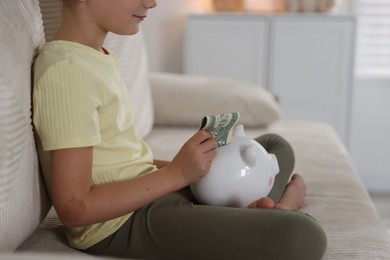 Photo of Little girl putting pocket money into piggy bank on sofa at home, closeup