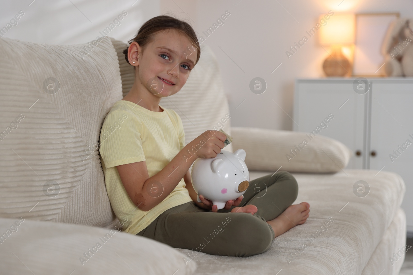 Photo of Little girl putting pocket money into piggy bank on sofa at home
