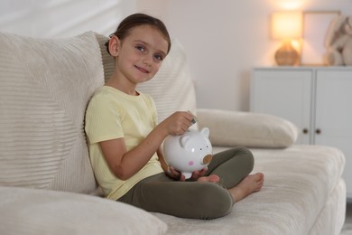 Photo of Little girl putting pocket money into piggy bank on sofa at home
