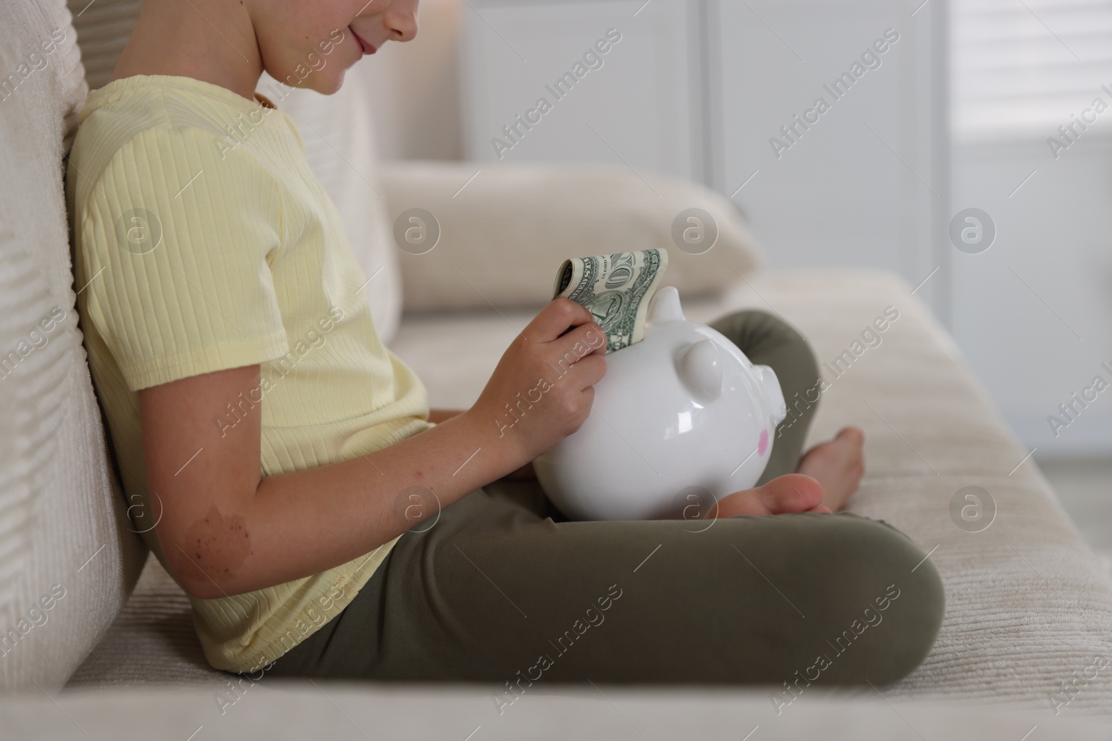 Photo of Little girl putting pocket money into piggy bank on sofa at home, closeup