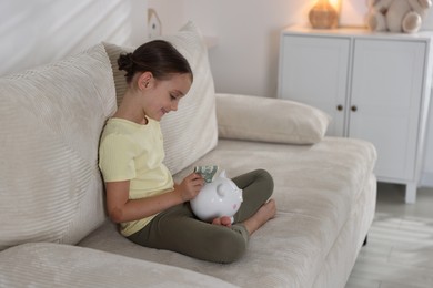 Photo of Little girl putting pocket money into piggy bank on sofa at home