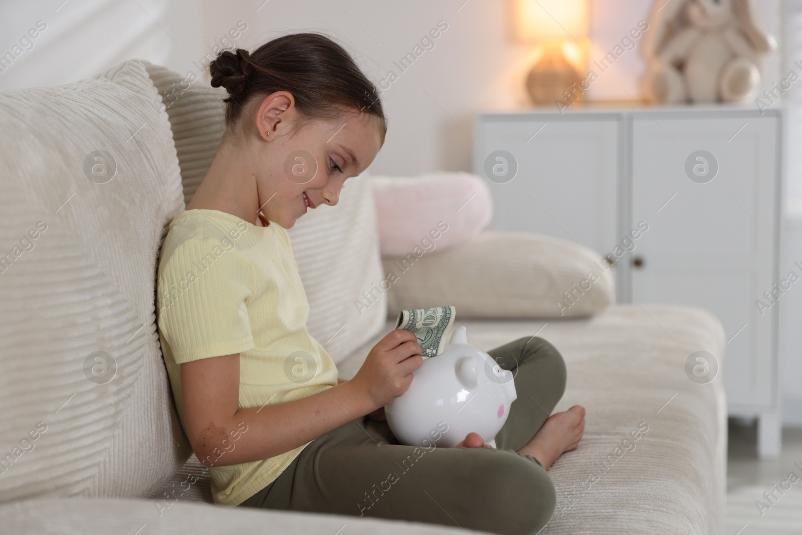 Photo of Little girl putting pocket money into piggy bank on sofa at home