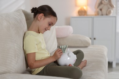 Photo of Little girl putting pocket money into piggy bank on sofa at home