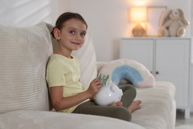 Photo of Little girl putting pocket money into piggy bank on sofa at home