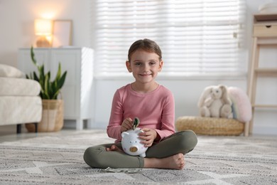 Photo of Little girl putting pocket money into piggy bank on floor at home