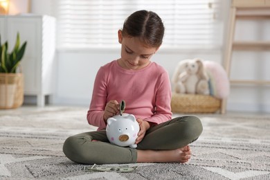 Photo of Little girl putting pocket money into piggy bank on floor at home
