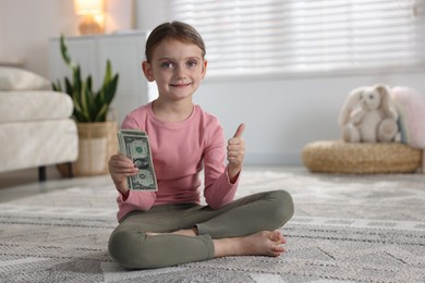 Photo of Little girl holding pocket money and showing thumbs up on floor at home