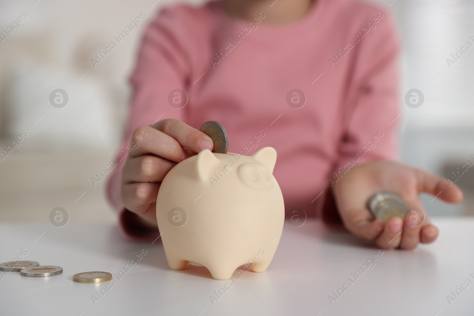 Photo of Little girl putting pocket money into piggy bank at white table indoors, closeup
