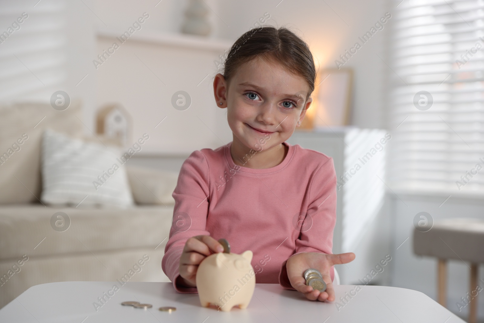 Photo of Little girl putting pocket money into piggy bank at white table indoors
