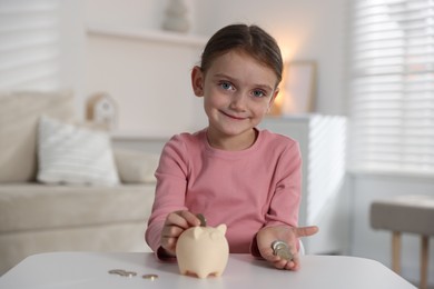 Photo of Little girl putting pocket money into piggy bank at white table indoors