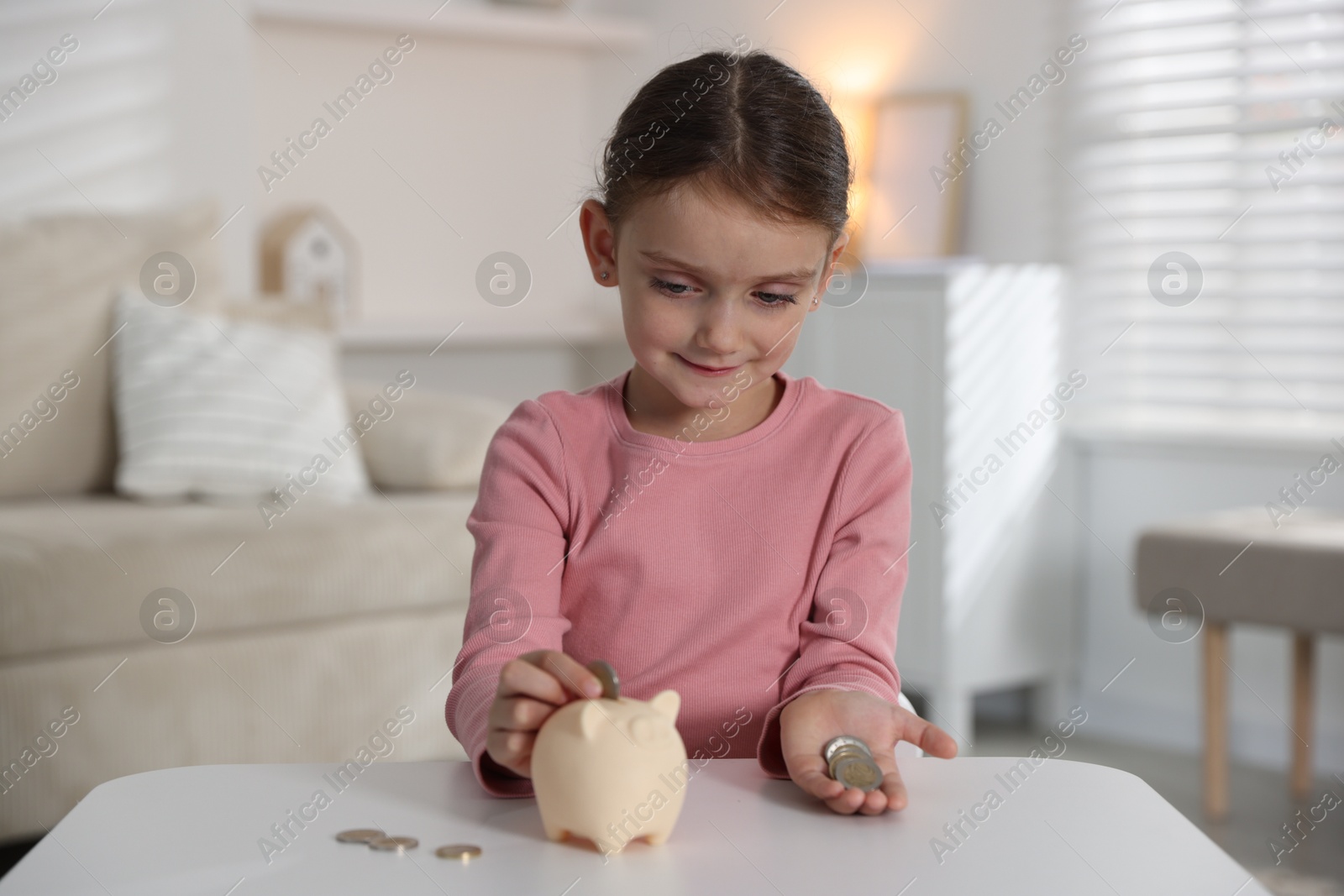 Photo of Little girl putting pocket money into piggy bank at white table indoors