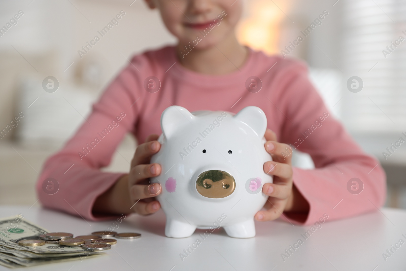Photo of Little girl with pocket money and piggy bank at white table indoors, closeup