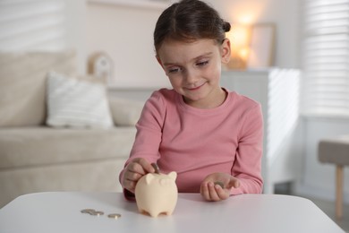 Photo of Little girl putting pocket money into piggy bank at white table indoors